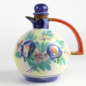 Könnte beinhalten: Ein runder Keramikkrug mit cremefarbener Basis, verziert mit handgemalten blauen und grünen Blumenmotiven. Er verfügt über einen leuchtend orangefarbenen Henkel, einen blauen Ausguss und einen passenden blauen Kugelstopfen mit Korkunterseite.