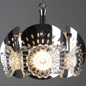 May include: A chrome-finished pendant light fixture with a unique design. The light features three clear, star-shaped crystal elements and a series of curved, perforated metal panels. The fixture hangs from a chrome chain and emits a warm glow.