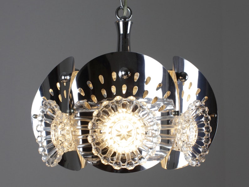 May include: A chrome-finished pendant light fixture with a unique design. The light features three clear, star-shaped crystal elements and a series of curved, perforated metal panels. The fixture hangs from a chrome chain and emits a warm glow.