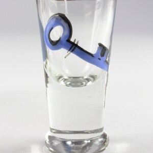 May include: Clear shot glass with a gold rim and a blue key design. The key is painted in a whimsical style with black accents. The glass is cylindrical with a slightly flared base.