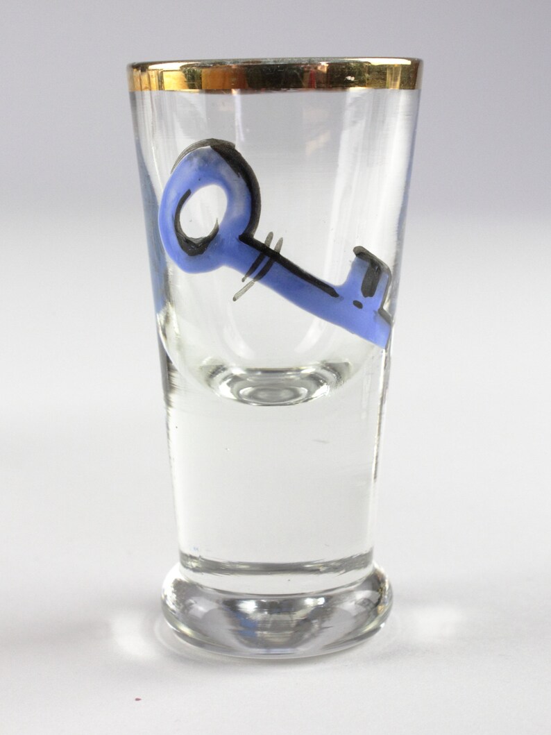 May include: Clear shot glass with a gold rim and a blue key design. The key is painted in a whimsical style with black accents. The glass is cylindrical with a slightly flared base.