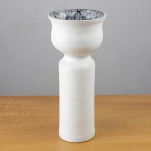Può includere: Un vaso alto in ceramica bianca con la parte superiore svasata e un interno a motivi. Il vaso ha una base cilindrica e una sezione centrale bulbosa. Il motivo interno è un mix di grigio e nero. Su una superficie di legno.