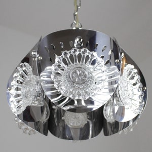 May include: A chrome-finished pendant light fixture with a flower-like design. The fixture features clear, decorative glass elements and a series of petal-shaped metal panels. The light hangs from a chain.