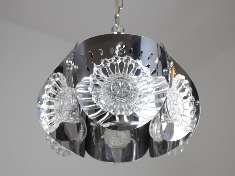May include: A chrome-finished pendant light fixture with a flower-like design. The fixture features clear, decorative glass elements and a series of petal-shaped metal panels. The light hangs from a chain.