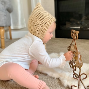 May include: A baby wearing a beige crocheted bonnet sits on a white rug and plays with a gold metal decorative stand.