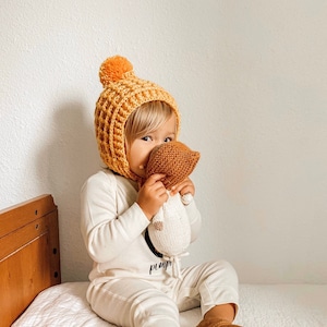 May include: A baby wearing a yellow crocheted hat with a pom pom and a white crocheted doll. The baby is sitting on a white bed with a wooden headboard.