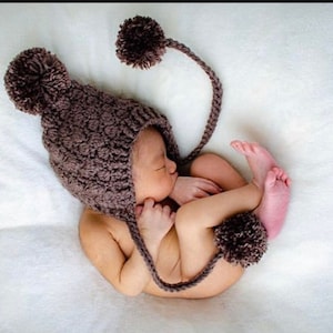May include: A brown knitted baby hat with two large pom-poms and a long tail. The hat is worn by a baby sleeping on a white blanket.