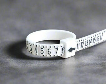 Könnte beinhalten: Ein weißer Kunststoff-Ringgrößenmesser mit schwarzen Zahlen von 2 bis 17. Der Größenmesser ist flexibel und kann verwendet werden, um den Umfang eines Fingers zu messen.