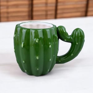 May include: A green ceramic mug shaped like a cactus, featuring a white interior and a handle designed to resemble a cactus arm. The mug has a glossy finish with small white dots, adding to the realistic cactus appearance. The mug is designed for beverages.