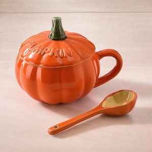 May include: An orange ceramic pumpkin-shaped mug with a lid and handle, and the word "PUMPKIN" embossed on the lid. A matching orange spoon with a yellow interior rests beside the mug. The mug's stem is green.