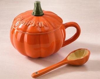 May include: An orange ceramic pumpkin-shaped mug with a lid and handle, and the word "PUMPKIN" embossed on the lid. A matching orange spoon with a yellow interior rests beside the mug. The mug's stem is green.