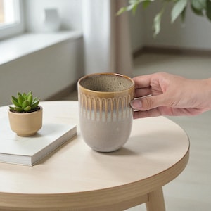 May include: A ceramic mug with a cylindrical shape, featuring a smooth, light beige base that transitions into a textured, vertical ribbed pattern near the rim, finished with a glossy, warm honey-brown glaze at the top edge and a speckled interior.
