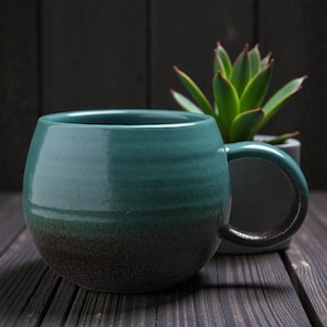 May include: A teal and dark brown glazed ceramic mug with a rounded body and a large handle. The mug is set on a dark wooden surface. A succulent plant in a grey pot is visible in the background. The mug's design features a colour gradient.