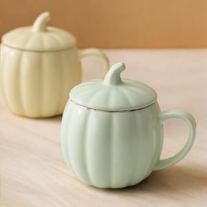 May include: Two ceramic mugs shaped like pumpkins, one pale yellow and the other light green. Each mug has a matching lid and a curved handle. The lids are trimmed with a silver-colored band. The mugs are on a light-colored wooden surface.