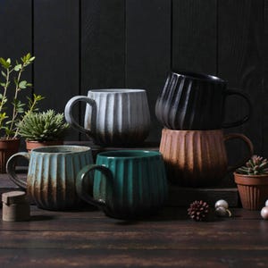 Puede incluir: Conjunto de seis tazas de cerámica acanaladas en varios colores, incluyendo turquesa, marrón, gris, negro y melocotón. Cada taza tiene forma redondeada y un asa. Las tazas están dispuestas sobre una superficie de madera oscura, con pequeñas plantas en macetas.