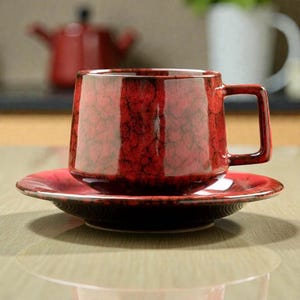 May include: A red ceramic coffee cup and saucer. The cup features a square handle and a textured, crackled glaze. The round saucer complements the cup's colour and finish. A red teapot is visible in the background.