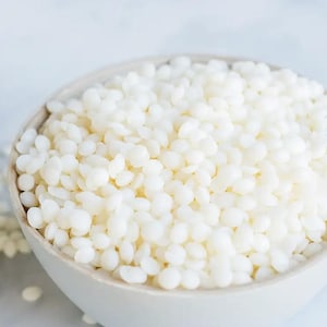 May include: A close-up shot of a white ceramic bowl filled with small, off-white, bead-like wax pellets. The pellets are densely packed, creating a textured surface. The bowl is set against a soft, blurred background.