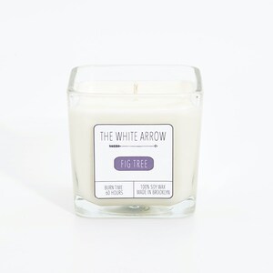 Puede incluir: Un tarro de vela cuadrado de vidrio lleno de cera blanca. La etiqueta dice "THE WHITE ARROW" y "FIG TREE". La etiqueta también indica "BURN TIME 60 HOURS" y "100% SOY WAX MADE IN BROOKLYN".