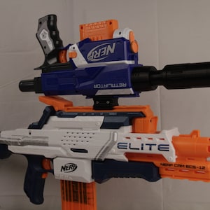 Op de afbeelding: Een witte en oranje Nerf Elite CAM ECS-12 blaster met een blauwe Nerf Retaliator blaster die bovenop is bevestigd. De Retaliator heeft een zwarte loopverlenging.
