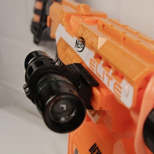 May include: A black camera mount attached to an orange Nerf Elite toy gun. The camera mount has a black camera lens and a green ring around the lens.
