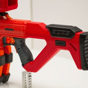 Op de afbeelding: Rood en zwart speelgoedpistool met een zwarte kolf en een zwarte greep. Het pistool heeft een magazijn aan de zijkant en een richtkijker bovenop. De kolf heeft de tekst "EDGE" erop gedrukt.