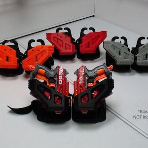 May include: Six 3D printed Nerf blaster holsters in orange, red, and grey. Two holsters are attached to a black leg strap with two Nerf blasters inserted. The text *Blasters NOT Included* is printed on the white surface.