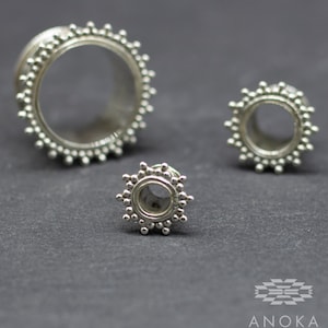 May include: Three silver ear gauges with a beaded design. The gauges are different sizes, with the largest gauge in the foreground.