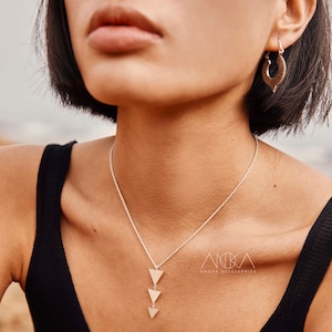 Puede incluir: Un collar de cadena de plata con tres colgantes triangulares que cuelgan de él. El collar lo lleva una persona con el pelo corto y oscuro. El texto "Anoka Accessories" es visible en la imagen.