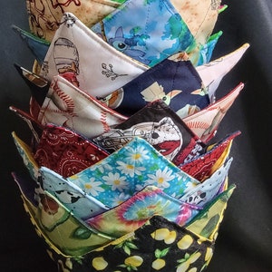 May include: A stack of handmade fabric bowl cozies with various patterns. Designs include lemons, daisies, tie-dye, and cartoon characters. The cozies are made of colorful fabric and are designed to hold bowls of hot food.