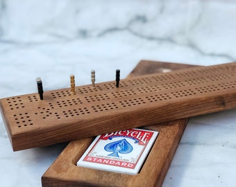 May include: A wooden cribbage board with a pack of cards inside. The board is stained brown and made of solid wood. It has a grid of holes for pegs and a scoring area.