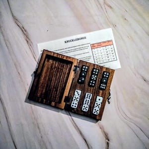 May include: A wooden box with a hinged lid, containing five dice. The box is open, revealing the dice inside. The box is labelled "Knucklebones" and has a chart with scoring rules.