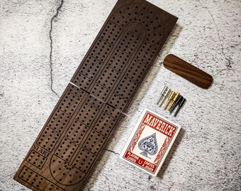 May include: A wooden cribbage board with a dark brown finish. The board is folded in half and has a start position marked on one side. The board is accompanied by a deck of playing cards and a set of metal pegs.