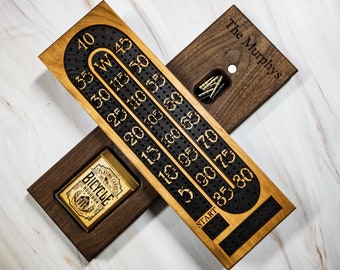May include: A wooden cribbage board with a black and white track for scoring points. The board has a personalized inscription "The Murphys" and a small compartment for holding playing cards. A deck of Bicycle playing cards is included.