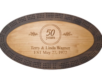 May include: A wooden oval-shaped cribbage board with a dark wood border and a light wood center. The center is engraved with "50 years", "Terry & Linda Wagner", and "EST May 27, 1972".