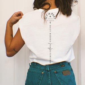 May include: White t-shirt with the text "SET APART SAVED BY GRACE" in a circular design and a vertical line of text that reads "REDEEMED RESTORED KINGDOM LIVING". The shirt is paired with blue jeans.
