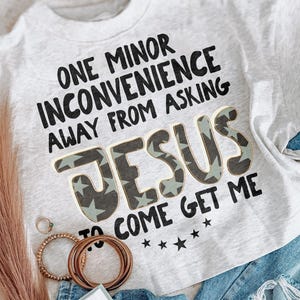 Pode incluir: Uma t-shirt cinza mesclado claro com texto preto que diz ONE MINOR INCONVENIENCE AWAY FROM ASKING JESUS TO COME GET ME. A palavra JESUS est&aacute; impressa em uma fonte grande e em negrito com um padr&atilde;o de estrelas verde escuro e claro.