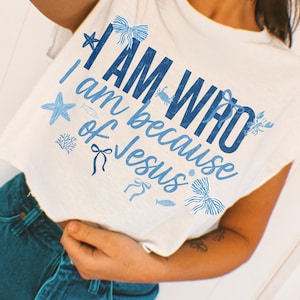 May include: A white short-sleeved t-shirt featuring blue text that reads I AM WHO I am because of Jesus. The design includes illustrations of a starfish, bows, a crab, a fish, and coral, all rendered in various shades of blue ink.