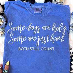 May include: A blue Comfort Colors t-shirt with white text that reads "Some days are holy. Some are just hard. Both still count." The shirt has a crew neck and a tag at the neckline. The shirt is surrounded by disco balls and a black and white checkered pattern.