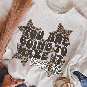 May include: A light gray sweatshirt with the text "YOU ARE GOING TO MAKE IT trust ME" in black and white. The words are set against a leopard print star design. The text "PSALM 23" is below the main text.