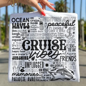 May include: White t-shirt with a black graphic design featuring the words "Cruise Vibes" and other cruise-related phrases and images, including a cruise ship, seashells, and a pineapple.