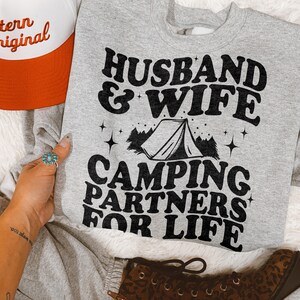 May include: A heather gray crewneck sweatshirt featuring black text that reads HUSBAND & WIFE CAMPING PARTNERS FOR LIFE, accompanied by a graphic of a tent, pine trees, and small stars. The sweatshirt is displayed alongside a partial orange and white cap.