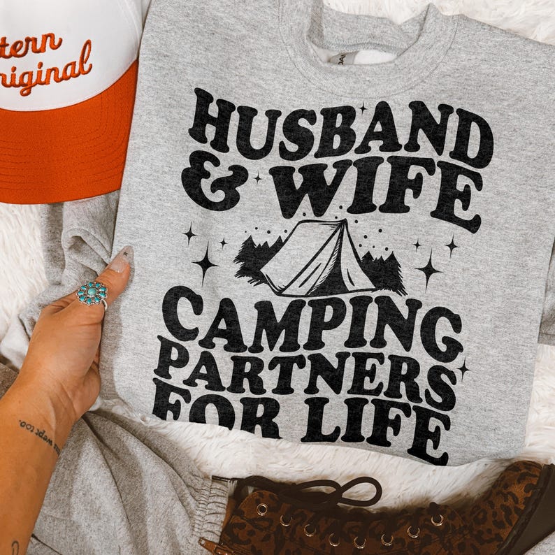 May include: A heather gray crewneck sweatshirt featuring black text that reads HUSBAND & WIFE CAMPING PARTNERS FOR LIFE, accompanied by a graphic of a tent, pine trees, and small stars. The sweatshirt is displayed alongside a partial orange and white cap.
