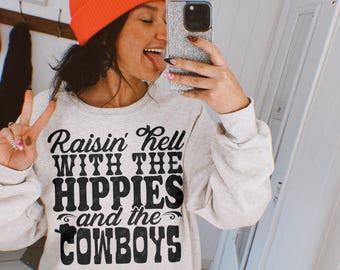 Op de afbeelding: Een grijs sweatshirt met de tekst "Raisin' Hell With The Hippies And The Cowboys" in zwart gedrukt.