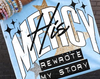 May include: Light blue t-shirt with the words "MERCY His" in white, with a gold glitter star behind the word "MERCY". The text "REWROTE MY STORY" is in a black rectangle. Black starbursts and a silver glitter star are also visible.