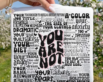 May include: A white t-shirt with the words "YOU ARE NOT" in a bold, retro font. The shirt is covered with smaller words like "anxiety," "stress," and "failure," creating a powerful message of self-acceptance and positivity.
