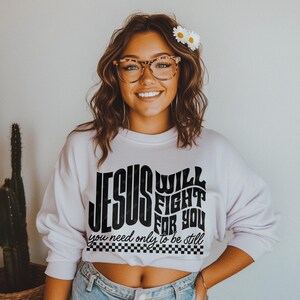 May include: A white sweatshirt with a black graphic print that reads "Jesus Will Fight For You, You Need Only To Be Still". The graphic is in a retro style font with a checkered border.