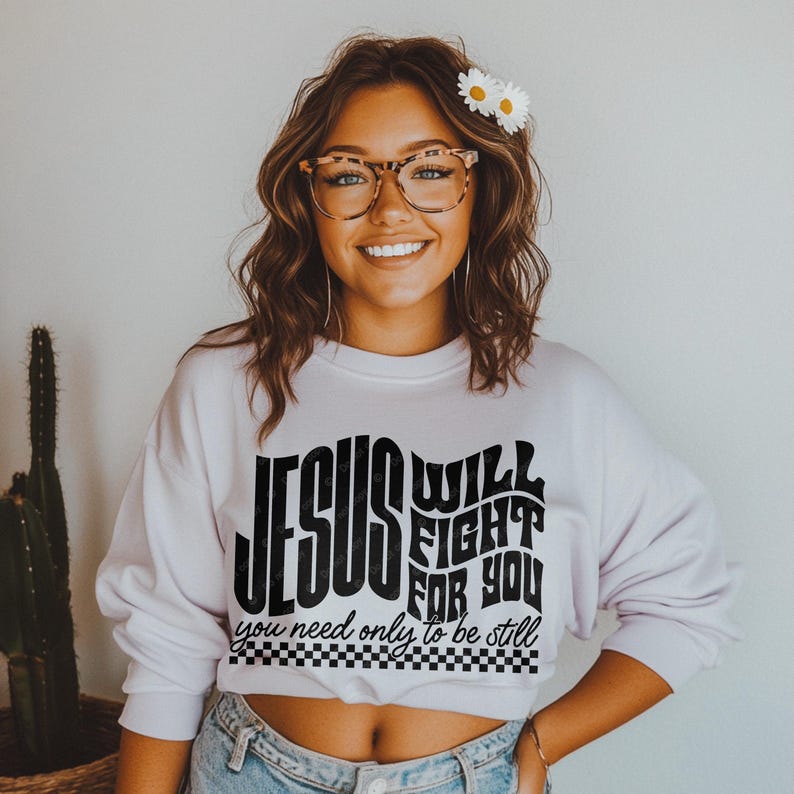 May include: A white sweatshirt with a black graphic print that reads "Jesus Will Fight For You, You Need Only To Be Still". The graphic is in a retro style font with a checkered border.
