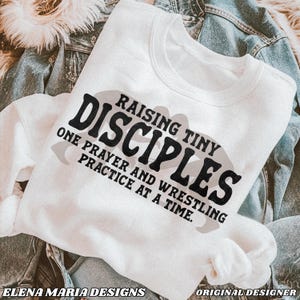 May include: White sweatshirt with black text that reads "Raising Tiny Disciples One Prayer and Wrestling Practice at a Time."