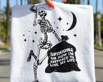 May include: White sweatshirt featuring a black skeleton graphic carrying a sack with the text "OPINIONS ON HOW I'M SUPPOSED TO LIVE MY LIFE." The design includes a crescent moon and stars.