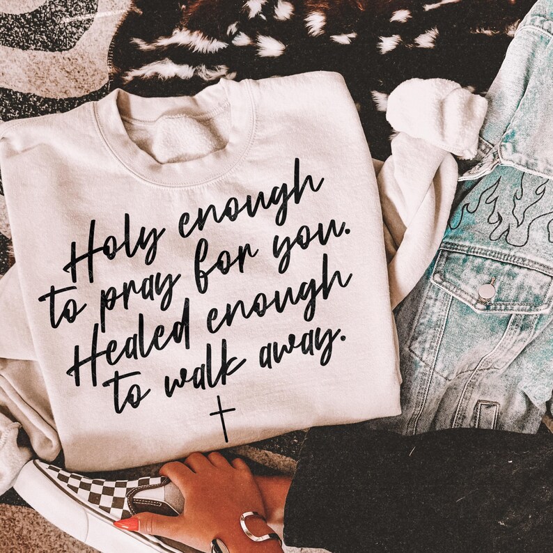 May include: Cream-colored sweatshirt with the text "Holy enough to pray for you. Healed enough to walk away." in black script, with a small cross below. A denim jacket and checkered slip-on shoe are visible.